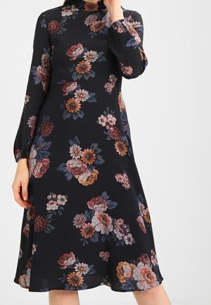 Robe midi noire à manches longues avec imprimé floral multicolore, col haut et jupe légèrement évasée, portée par une personne avec une main près du visage.