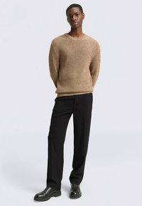 Bēša ribota adījuma džemperis ar apaļu kakla izgriezumu, kombinēts ar melnām piegrieztām biksēm un melniem zābakiem, radot ikdienišķu, taču pārdomātu izskatu.