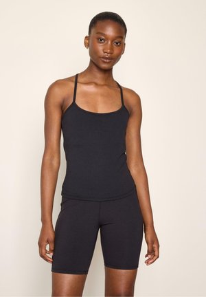 Kvinde iført en sort tætsiddende tanktop med tynde stropper og matchende sorte cykelshorts, stående mod en ensfarvet lys baggrund.