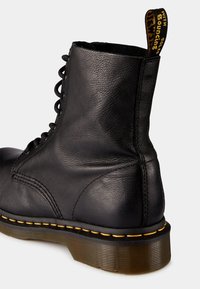 Bota de cuero negra con tobillo alto, puntera redonda y diseño de cordones. Presenta un detalle de costura amarilla en la suela y una lengüeta.