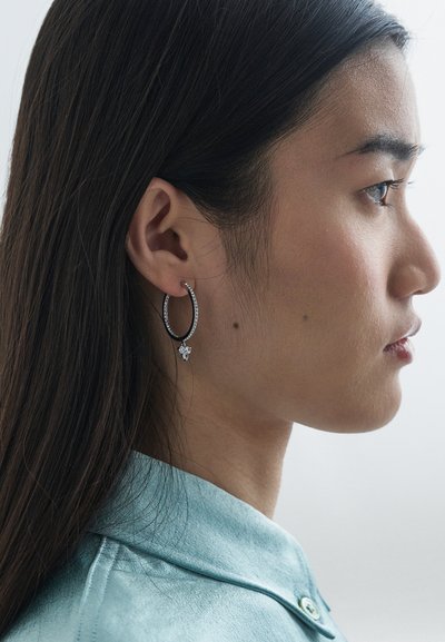 Mlada ženska s dolgimi temnimi lasmi, oblečena v svetlo modro majico in z velikimi srebrnimi uhani v obliki obroča, na katerih je majhen cvetlični obesek.