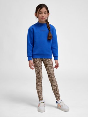 Ung pige står med langt flettet hår, iført en blå sweatshirt, leopardmønstrede leggings og hvide sneakers med orange detaljer.