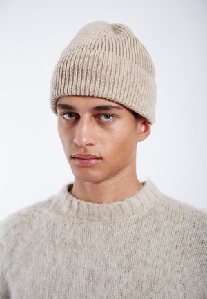 Young man with light skin wearing a beige knit beanie and a fuzzy beige sweater, looking directly ahead against a plain background.