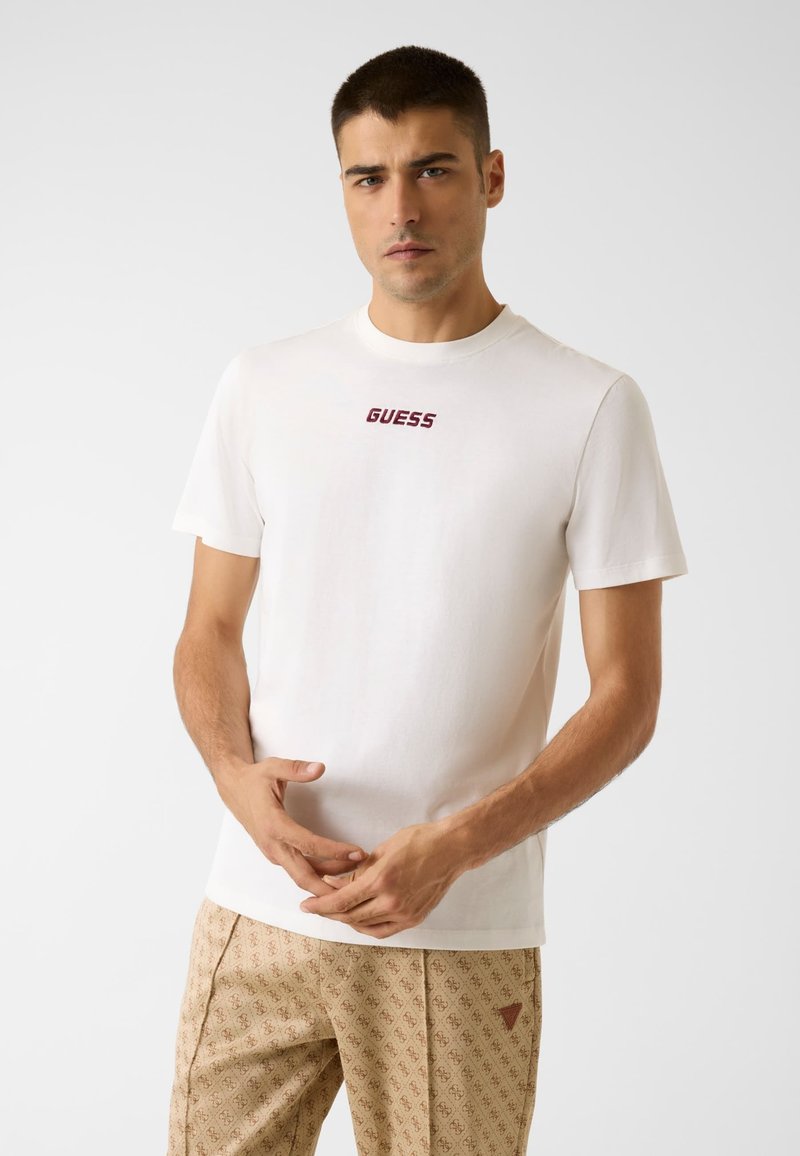 Biały bawełniany t-shirt z bordowym logo "GUESS" na piersi, okrągłym dekoltem i krótkimi rękawami, zestawiony z beżowymi spodniami w wzory.