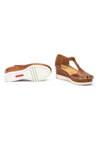 Scarpe a zeppa in pelle marrone con design perforato, cinturino a T e suola in gomma bianca. Dettaglio di cucitura notevole lungo il bordo.