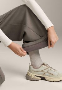 Šedé teplákové nohavice s textúrovaným povrchom, s nastaviteľným manžetom. Svetlobežové tenisky s detailmi z priedušnej sieťoviny a hrubou podrážkou, nosené s rebrovanými ponožkami.