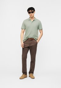 Άνδρας που φοράει πράσινο πόλο, καφέ παντελόνι με ζώνη, μπεζ αθλητικά παπούτσια και μαύρα γυαλιά ηλίου, στέκεται μπροστά σε απλό λευκό φόντο.