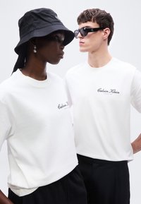 Δύο μοντέλα φορούν λευκά T-shirts Calvin Klein· το ένα φοράει μαύρο καπέλο τύπου bucket και σκουλαρίκια, το άλλο μαύρα γυαλιά ηλίου.