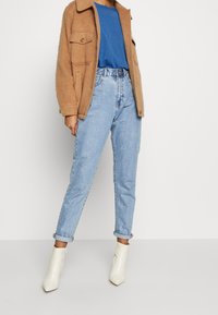 Hnědá bunda z vlněné směsi s předními kapsami, modré tričko s kulatým výstřihem, světle modré džíny a bílé špičaté kotníkové boty. Otočené manžety džín.