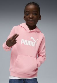 Ružová mikina s kapucňou a bielym logom "PUMA" a grafikou skákajúcej mačky. Mäkkučký materiál, predná vrecko, rebrované manžety a lem. Používateľ ukazuje znak pokoja.