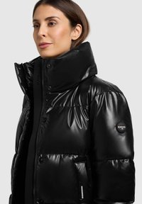 Doudoune noire matelassée avec un col montant, finition brillante, fermeture éclair et boutons pression, dotée d'une poche latérale et d'un écusson logo sur la manche.