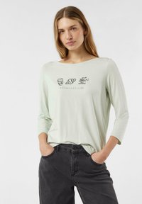 Jasnozielona koszula z długim rękawem, z dekoltem w serek i prostymi ilustracjami nawiązującymi do śniadania na froncie, zestawiona z ciemnymi dżinsami.