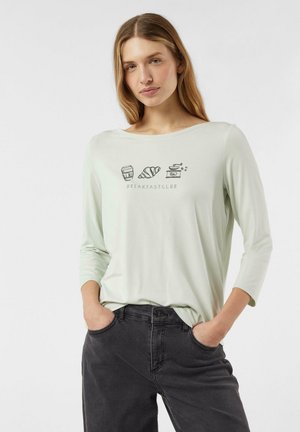 Jasnozielona koszula z długim rękawem, z dekoltem w serek i prostymi ilustracjami nawiązującymi do śniadania na froncie, zestawiona z ciemnymi dżinsami.