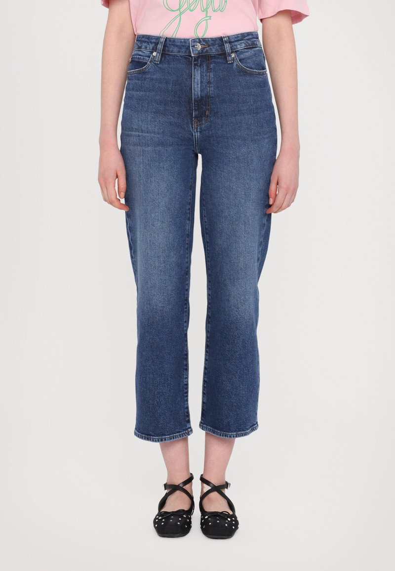 High-waisted blue denim jeans with a cropped leg cut, featuring subtle fading and a classic 5-pocket design. Paired with black strappy shoes.