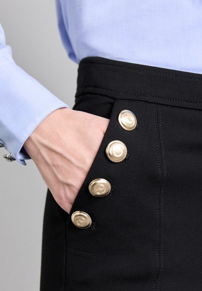 Black skirt with gold buttons on the side, featuring a structured design. Smooth fabric texture and visible stitching detail.