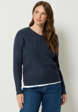 Femme aux longs cheveux bruns portant un pull bleu foncé en maille torsadée à col en V par-dessus une chemise blanche et un jean bleu, debout devant un fond uni.