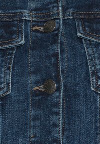 Tumma sininen farkkukangas, jossa on kaksi rintataskua mustilla napituksilla, näkyvillä saumoilla ja hieman kuluneella pinnalla.