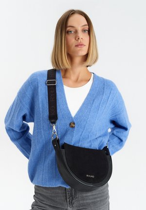 Borsa a tracolla in pelle nera e suede a forma di mezzaluna, con una tracolla staccabile con stampa del logo. Il modello indossa un cardigan blu.