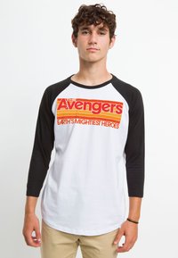 Weißes Raglan-T-Shirt mit schwarzen Ärmeln, auf der Vorderseite mit einer bunten "Avengers"-Grafik. Aus Baumwollmaterial, figurbetonter Schnitt.