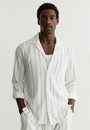 Mand i hvid, let knap-op-skjorte med tynde, lodrette sorte striber, hvid undertrøje, matchende stribede bukser, hænder i lommerne.