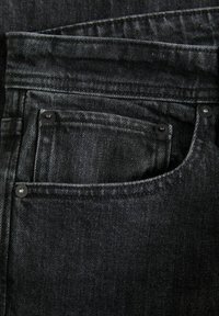 Jeans de mezclilla negra con un primer plano del bolsillo delantero, que presenta remaches de metal, tela texturizada y detalles de costura visibles.