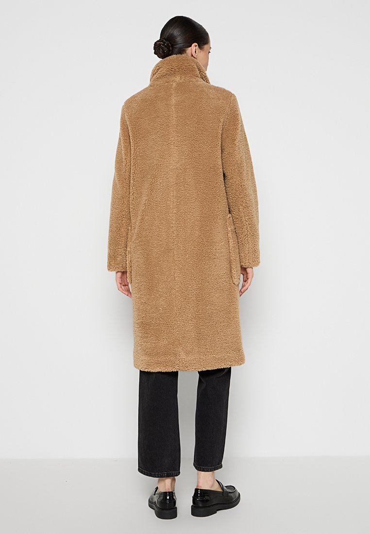 Manteau en shearling marron avec un col haut, des manches longues et des poches latérales, associé à un jean noir et des chaussures noires brillantes.