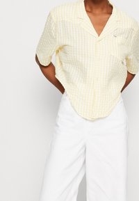 Chemise à manches courtes en vichy jaune avec un col, une poche avant et une coupe décontractée, associée à un pantalon blanc.