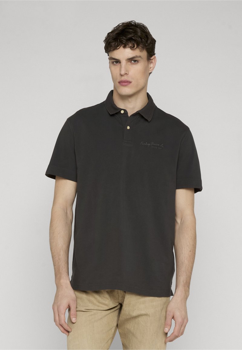 Jeune homme aux cheveux courts et bouclés portant un polo noir et un pantalon beige, debout devant un fond clair uni.
