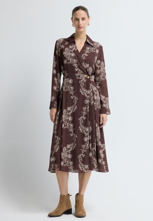 Langärmliges Wickelkleid in sattem Burgunderrot mit cremefarbenem Paisleymuster, ausgestattet mit einem Kragen, Knopfverschluss und fließendem Rock. Getragen mit beigefarbenen Stiefeletten.