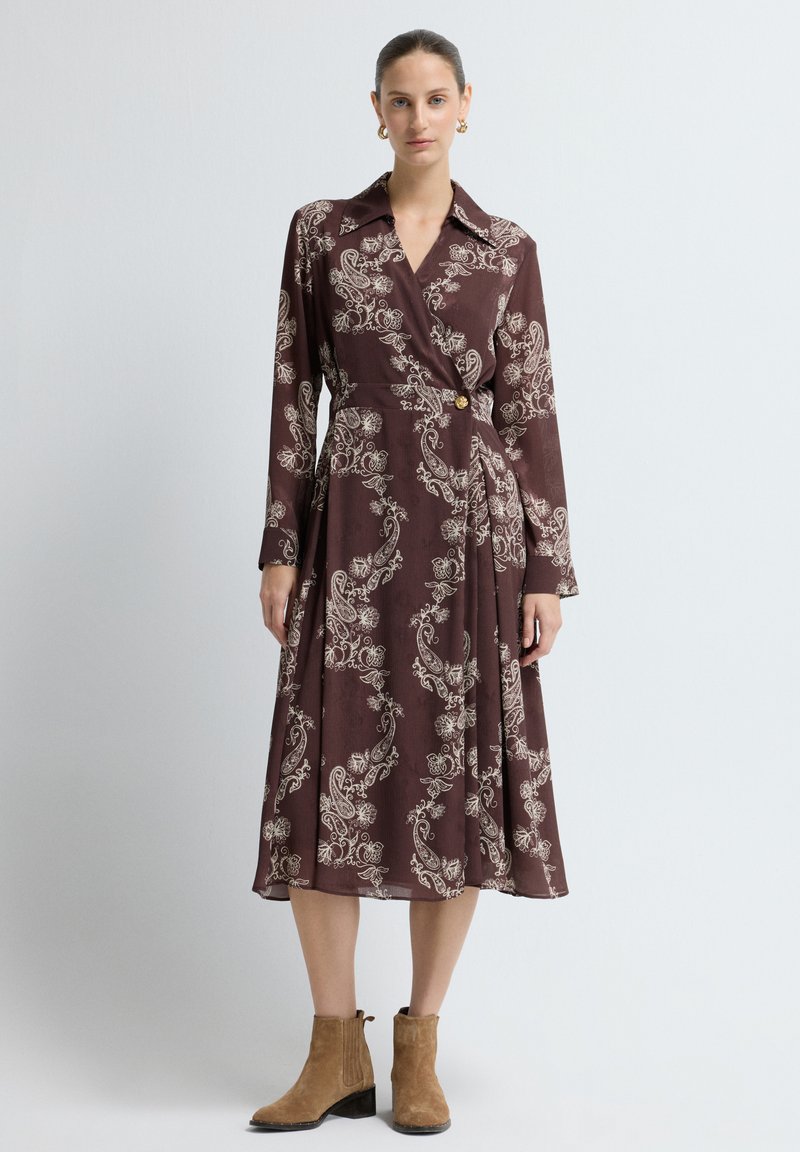 Langärmliges Wickelkleid in sattem Burgunderrot mit cremefarbenem Paisleymuster, ausgestattet mit einem Kragen, Knopfverschluss und fließendem Rock. Getragen mit beigefarbenen Stiefeletten.