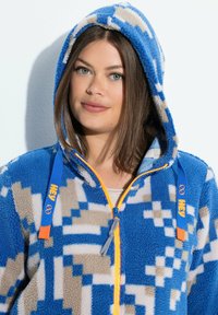 Giovane donna che indossa un hoodie in pile con motivo blu, beige e bianco, con zip gialla e lacci blu etichettati "HEY", appoggiata a un muro bianco.