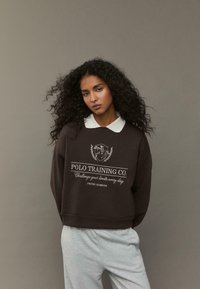 Felpa marrone con colletto bianco, stampa grafica con testo e orlo a costine. Indossata con pantaloni della tuta grigi chiari. Sfondo semplice.