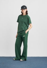 Πράσινο βαμβακερό t-shirt με μικρό λευκό λογότυπο Nike, συνδυασμένο με φαρδιά πράσινη φόρμα με μπλε γραφικό "Nike". Μαύρο καπέλο στο κεφάλι.