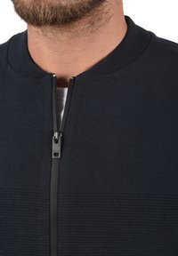 Tamsiai mėlynas zip-up džemperis, pasižymintis tekstūruotu dizainu, raukšlėtu apykakle ir lygiu užtrauktuku. Minimalistinis stilius be raštų.
