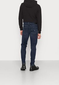 Jeans de mezclilla azul oscuro con ajuste slim, que cuentan con bolsillos traseros y una etiqueta de marca. Combinados con una sudadera corta negra y botas negras.