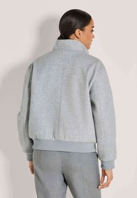 Frau mit dunkel gebundenem Haar, die eine hellgraue hochgeschlossene Jacke und passende graue Hosen trägt, von hinten vor einem schlichten Hintergrund gezeigt.