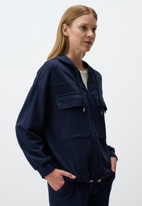Hoodie zippé marine avec deux poches avant, capuche à cordon de serrage et accents en métal doré. Tissu doux avec une coupe décontractée et des poignets côtelés.