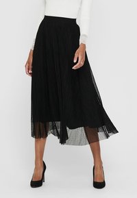 Jupe midi plissée noire avec une large ceinture, texture aérienne et une couche transparente, associée à des chaussures à talons hauts noires.