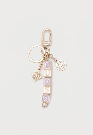 Portachiavi color oro con blocchi lettere viola e bianchi che formano la scritta "GUESS" e due ciondoli circolari decorativi attaccati a un moschettone.