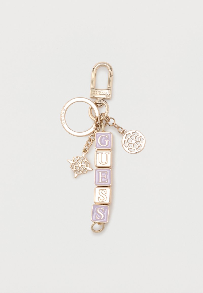 Μπρελόκ σε χρυσό τόνο με μωβ και λευκά γράμματα που σχηματίζουν τη λέξη "GUESS" και δύο διακοσμητικά κυκλικά γούρια προσαρτημένα σε μια παρωστήρα.