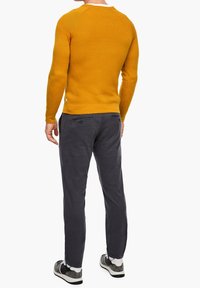 Gelber Strickpullover mit langen Ärmeln, Rundhalsausschnitt und strukturiertem Stoff; kombiniert mit dunkelgrauen Hosen und grauen Schuhen.