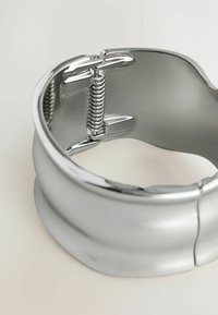 Hopeinen metallinen rannekoru, jossa on sileä pinta, varustettu jousimekanismilla ja kiillotetulla, heijastavalla pinnalla. Ei näkyvää kuviointia.