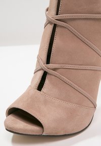 Beige ruskind højhælede sko med åben tå, med krydsede stropper og et udskæringsdesign langs siden. Glat, struktureret overflade.