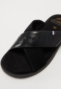 Svart sklisandal med en bred lærøvre med preget "bugatti"-logo, myk semsket tekstur og en gummisåle med en subtil glans.