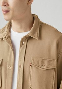 Beige, gestructureerde overhemdjas met een kraag, frontale knoopsluiting en twee borstzakken. Voorzien van een rood accentlabel. Witte T-shirt zichtbaar eronder.