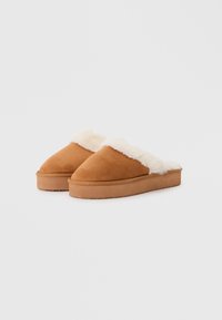 Pantofole in suede marrone con un morbido colletto color crema, punta arrotondata e suola piatta in gomma, progettate per il comfort e la facilità d'uso.