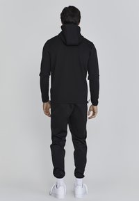 Zwarte hoodie met een hoge kraag en een getailleerd ontwerp, gecombineerd met bijpassende joggers. Het materiaal lijkt soepel en rekbaar. Witte sneakers maken de look compleet.