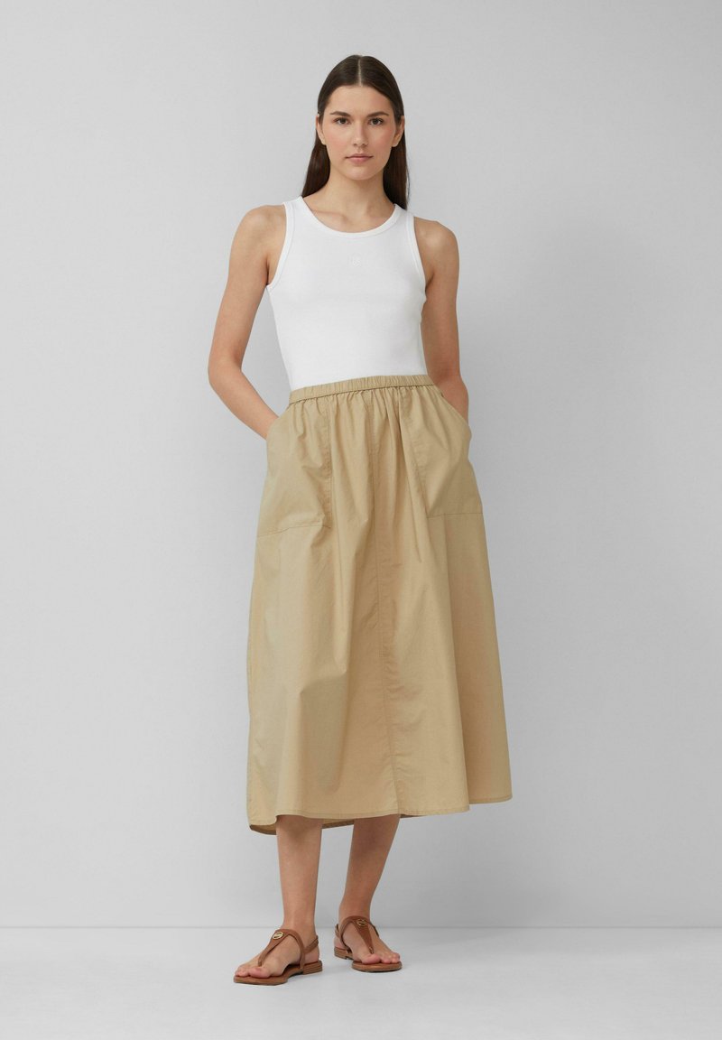 Beige, midi-jurk met een elastische tailleband en zijzakken, gecombineerd met een witte tanktop en bruine sandalen. Soepele, lichte stof.