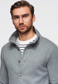 Šedý fleecový pulovr s vysokým límcem, vybavený dvěma knoflíky a kontrastní proužkovanou košilí pod ním. Viditelné jsou také brýle a vousy.