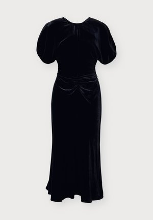 Femme en robe midi en velours noir avec manches courtes bouffantes et taille froncée, portant des bottines noires à talons hauts, debout sur fond blanc.